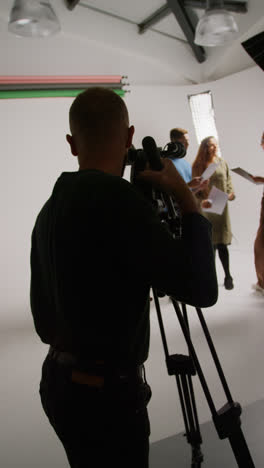 Vertical-Video-Of-Film-Camera-Operator-Shooting-Director-Talking-With-Male-And-Female-Actors-Holding-Scripts-Rehearsing-For-Shooting-Movie-Or-Video-In-Studio